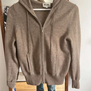 Beige kashmirhoodie från Soft Goat - Mysig beige hoodie från Soft Goat i kashmir. Tröjan har dragkedja framtill, huva med snörning och ribbade muddar. Perfekt för dig som gillar en clean och stilren look med lyxig känsla. Liten defekt längst ner på dragkedjan där den har lossnat lite från sömmen men annars fint skick