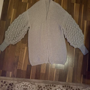 Beige stickad kofta med puffärmar - Superfin beige stickad kofta med breda puffärmar och coolt bubbelmönster på ärmarna. Koftan har en öppen front och ribbade muddar vid ärmslut och nederkant. Perfekt för lager-på-lager och ger en trendig vibe till din outfit.