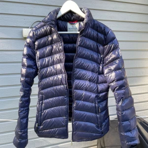 Mörkblå Moncler dunjacka - Säljer en mörkblå dunjacka från Moncler med glansig finish och klassisk quiltad design. Jackan har dragkedja framtill, ståkrage och Moncler-logga på ärmen. Insidan har ett unikt serietryck. Perfekt för kalla dagar och riktigt snygg till streetwear. Priset är inte skrivet i sten!