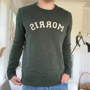 Grön stickad tröja från Morris - Mörkgrön stickad tröja från Morris med rund halsringning och vit broderad logga framtill. Tröjan har ribbade muddar vid ärmslut och nederkant, perfekt för dig som gillar klassisk och stilren look.