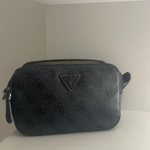 Svart väska från Guess - Snygg svart axelväska från Guess med diskret mönster och metalllogga framtill. Väskan har två fack med dragkedja och ljus insida med Guess-tryck. Perfekt storlek för mobil, plånbok och småsaker. 