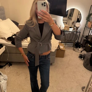 Grå kavaj med bälte och croppad passform - Säljer en stilren grå kavaj med croppad passform och bälte i midjan med metallring. Jackan har trekvartsärmar och en snygg, minimalistisk look som passar perfekt till jeans eller kjol.