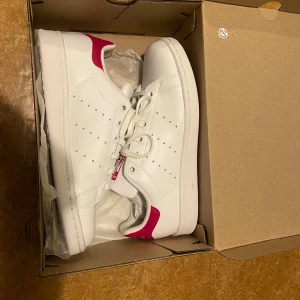 Adidas Stan Smith  - Helt oanvända med alla etiketter kvar (fel köpt, gick inte att lämna tillbaka) Adidas Stan Smith sneakers i vitt med snygga rosa detaljer på häl och plös. Klassisk låg modell med snörning. Storlek 37 1/3. Köptes för 800 kr.