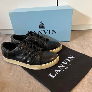 Svarta Lanvin sneakers i skinn - Snygga svarta sneakers från Lanvin i äkta skinn med rund tå och vita gummisulor. Klassisk design med svarta snören och metallögla med Lanvin-logga. Perfekta för dig som gillar stilrena och exklusiva sneakers med premiumkänsla.