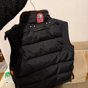Svart dunväst från Parajumpers - Snygg svart dunväst från Parajumpers med fluffig brun krage i fuskpäls, orange detaljer på sidorna och flera praktiska fickor med dragkedja och knappar. Västen har quiltad design och grov dragkedja framtill. Perfekt för kalla dagar när du vill se cool ut.