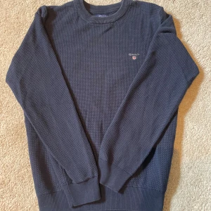 Mörkblå tröja från GANT i bomull - Mörkblå långärmad tröja från GANT med diskret logga på bröstet. Tröjan har en strukturerad, småmönstrad yta och ribbade muddar vid ärmslut och nederkant. Tillverkad i mjuk bomullsmix som känns skön mot huden.