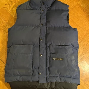 Mörkblå dunväst från Canada Goose - Snygg äkta mörkblå vintage dunväst från Canada Goose med två stora fickor framtill och logga på ena fickan. Västen har krage och knappar. Perfekt för lager-på-lager och en clean look.