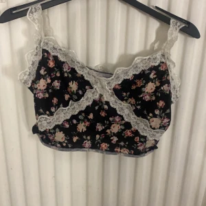 Blommig linnetopp med spets - Supersöt linnetopp med svart botten och blommigt mönster i rosa, lila och grönt. Toppen har vita spetsdetaljer längs kanterna och axelbanden, samt en omlott-look framtill. Perfekt för dig som gillar romantisk och feminin stil.