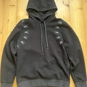 Svart Neil Barrett hoodie, strl L - Svart hoodie från Neil Barrett med ikoniska blixtar i grått/svart över axlarna. Tillverkad i en mjuk bomullsblandning med viskos och elastan för extra komfort. Perfekt för dig som gillar streetstyle med edge. Banden är lite noppriga, i övrigt fint skick. 