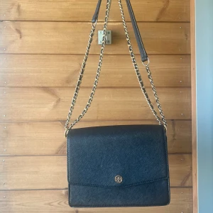 Svart axelväska från Tory Burch - Snygg svart axelväska från Tory Burch i skinn med guldfärgade kedjedetaljer. Väskan har en stilren, fyrkantig form och ett litet logomärke framtill. Perfekt storlek för det viktigaste och en klassisk look som passar till många outfits.