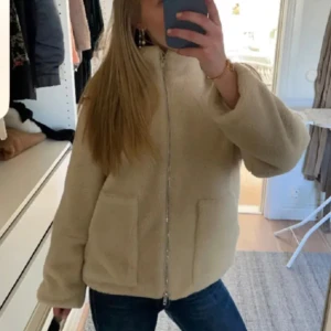 Beige teddyjacka  - Mysig beige teddyjacka med hög krage och två stora fickor framtill. Jackan har en rak passform och stängs med dragkedja. Perfekt för dig som gillar en chill och trendig look.