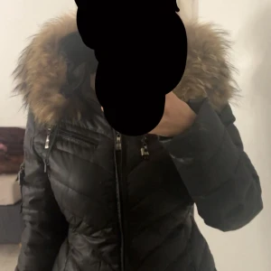 Svart pufferjacka med fuskpäls - org pris 3500 Snygg svart pufferjacka med stor huva kantad av brun fuskpäls. Jackan har dragkedja framtill, flera fickor med dragkedjor och markerad midja. Perfekt för kalla dagar när du vill hålla stilen och värmen.