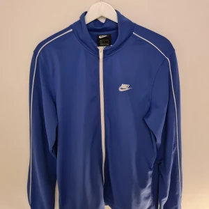 Blå Nike ziptröja med vita detaljer M - Sportig blå ziptröja från Nike med vita ränder längs ärmarna och vit dragkedja. Klassisk logga på bröstet och ståkrage. Tröjan har fickor i sidorna och är tillverkad i ett mjukt, lätt polyestermaterial. Perfekt för träning eller chill.