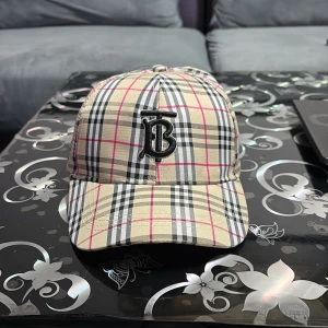 Burberry rutig keps i bomull - Snygg keps från Burberry i klassiskt rutigt mönster med beige, svart, vitt och rosa detaljer.Säljer p.g.a att jag inte använder den.