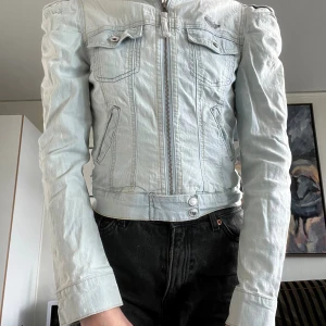Ljusblå jeansjacka med puffärmar - Säljer en ljusblå jeansjacka från Grunt Girl med markerade axlar och coola detaljer. Jackan har dragkedja framtill, två bröstfickor med lock och knappar samt två snedställda fickor. Croppad modell med lång ärm och snygg passform.
