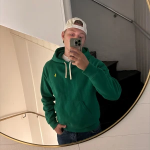 Grön hoodie från Polo Ralph Lauren - Säljer en grön hoodie från Polo Ralph Lauren med klassisk gul logga på bröstet. Tröjan har huva med vita snören och en stor magficka. Perfekt för dig som gillar en avslappnad och stilren look.
