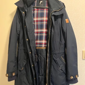 Marinblå parkas från Five Seasons - Snygg marinblå parkas från Five Seasons med rutigt foder i rött, blått och vitt. Jackan har huva, dragkedja och knappar framtill, samt praktiska fickor och muddar vid ärmslut. Perfekt för kyliga dagar och har en klassisk, tidlös look.