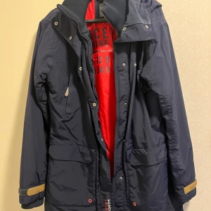 Mörkblå parkas från Race Marine - Snygg mörkblå parkas från Race Marine med röd insida, huva och stora fickor framtill. Jackan har dragkedja och knappar, samt justerbara ärmslut med kardborre. Perfekt för kyliga dagar och har en sportig vibe.