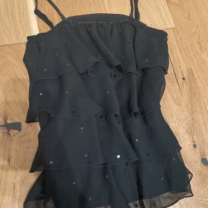 Svart volangklänning med paljetter Lindex - Svart kortklänning från Lindex med tunna axelband och flera lager volanger. Klänningen är dekorerad med små svarta paljetter både längs kanten och utspritt över hela tyget. Perfekt för dig som gillar en glittrig och lekfull stil.