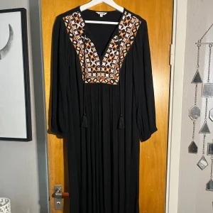 Svart långklänning broderad  - Svart långklänning från Indiska med färgglada broderier i orange, rosa, vitt och gult framtill. Klänningen har v-ringning med snörning och långa ärmar. Tyg i mjuk, lätt kvalitet och bohemisk känsla. Perfekt för dig som gillar unika detaljer och mönster.Storleken är XS men passar upp till M.