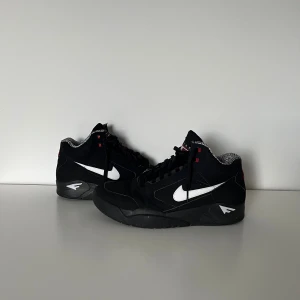 Nike Air Flight svart/vit sneakers - Säljer ett par svarta Nike Air Flight sneakers med vita swoosh-loggor och röda detaljer. Skorna har högt skaft, snörning och mönstrat innerfoder. Ovandelen är i mocka och syntet, sulan är svart och tåformen är rund. Perfekta för dig som gillar klassisk basketstil.
