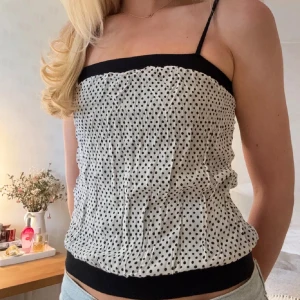 Fest topp - Supersöt linnetopp med tunna axelband och svart kant upptill och nertill. Toppen är vit med små svarta prickar och har en lätt rynkad effekt i tyget. Perfekt till jeans eller kjol för en trendig look.