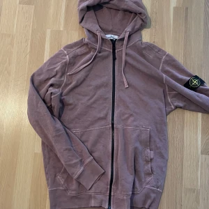 Ljuslila hoodie från Stone Island - Säljer en ljuslila hoodie från Stone Island med dragkedja och huva. Tröjan har den klassiska loggan på ärmen och stora fickor framtill. Perfekt för dig som gillar streetwear och vill ha något bekvämt och snyggt.
