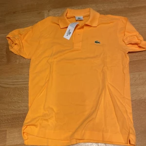 Lacoste - Orange Original pikétröja - Ny - Snygg orange pikétröja från Lacoste med klassisk krage och korta ärmar. Tröjan har det ikoniska gröna krokodilmärket broderat på bröstet och är gjord i bomull. Perfekt för dig som vill ha en fräsch och sportig look.
