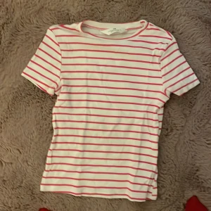 Vit och rosa randig t-shirt från H&M - Vit t-shirt från H&M med tunna rosa ränder och ribbad struktur. Klassisk rund halsringning och korta ärmar. Perfekt för dig som gillar en enkel men ändå snygg stil. Gjord i mjuk bomull som känns skön mot huden.