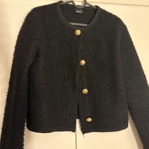 Svart cardigan jacka från Gina Tricot - Svart cardigan jacka från Gina Tricot i storlek XS. Jackan har en fluffig, stickad yta och är dekorerad med fyra snygga guldfärgade knappar framtill. Rund halsringning och långärmad modell, perfekt för lager-på-lager-stil. Old money style