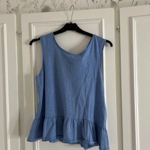 Blå ärmlös linnetopp med volang - Säljer en ljusblå ärmlös linnetopp med volangkant nedtill och snygg knytning i ryggen. Toppen har rund halsringning och är gjord i mjukt bomullstyg.