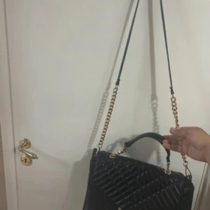 Svart quiltad axelväska med guldkedja - Snygg svart axelväska med quiltad design och guldfärgade kedjedetaljer. Väskan har både handtag och lång axelrem, perfekt för att bära på olika sätt. Stängs med lock och har en stilren look som lyfter varje outfit.