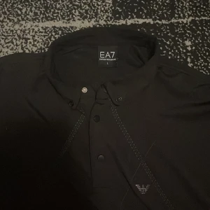 Svart EA7 kortärmad henley - Svart henley från EA7 Emporio Armani med korta ärmar och knappslå framtill. Skjortan har diskret EA7-logga på bröstet och stilrena sömmar som ger en clean look. Perfekt för dig som gillar snygga och enkla plagg med premiumkänsla.