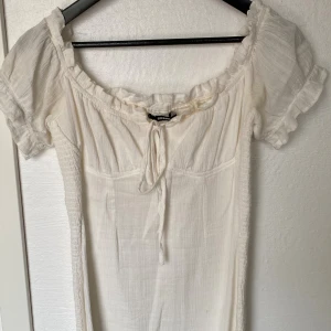 Vit kortklänning från BikBok - Supersöt vit kortklänning från BikBok med puffärmar, smockdetaljer och knyt framtill. Klänningen har en lätt rynkad struktur och offshoulder-krage med volangkant. Perfekt för varma dagar och ger en romantisk vibe.