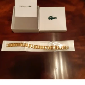 Guldfärgad länkarmband från Lacoste - Säljer ett stilrent guldfärgat länkarmband från Lacoste. Armbandet har en klassisk design med breda länkar och kommer i originalförpackning med Lacoste-logga. Perfekt för dig som vill uppgradera din klocka med en snygg accessoar.