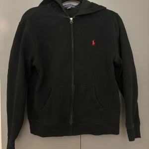 Svart hoodie från Polo Ralph Lauren - Svart hoodie från Polo Ralph Lauren med dragkedja och huva. Klassisk design med röd broderad logga på bröstet och känguruficka framtill. Perfekt för en chill och stilren look.
