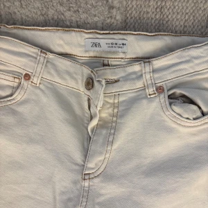 Vita baggy/biotcut jeans - Snygga vita jeans från Zara med bootcut passform och klassisk femficksdesign. Byxorna har ljusbruna kontrastsömmar, dragkedja och knapp framtill samt slitsdetaljer vid bensluten. Perfekta för dig som gillar en clean och fräsch look.Tveka inte på att höra av dig vid frågor eller intresse!
