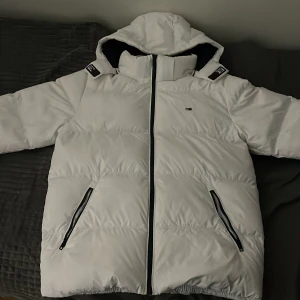 Vit pufferjacka från Tommy Jeans XL - Säljer en fet vit pufferjacka från Tommy Jeans i storlek XL. Jackan har huva, svarta detaljer vid dragkedjor och fickor, samt ett litet Tommy Jeans-logo på bröstet. Insidan är mörkblå med röd och vit detalj upptill. Perfekt för kalla dagar. 
