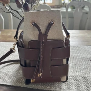 Brun och beige axelväska med snörning - Snygg axelväska i bucket-modell med brunt skinn och beige canvas. Väskan har justerbar axelrem, cool snörning upptill och detaljer i guld. Perfekt storlek för det viktigaste och ger en trendig touch till din outfit.