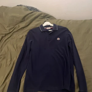 Mörkblå långärmad pikétröja Moncler - Snygg mörkblå långärmad pikétröja från Moncler med klassisk krage och diskret vit rand på kragen. Tröjan har broderad Moncler-logga på bröstet och knäppning framtill. Slim fit-modell i bomull som passar perfekt till jeans eller chinos.