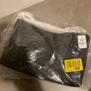 Svart axelväska från Calvin Klein - Stilren svart axelväska från Calvin Klein med minimalistisk design och silverfärgad logga framtill. Väskan har en mjuk, slät yta och dragkedja upptill. Perfekt för dig som gillar enkel och klassisk stil.Kvitto finns ej men självklart äkta