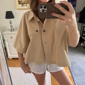 Beige overshirt med kort ärm - Stilren beige overshirt med korta, vida ärmar och klassisk krage. Skjortan har knappar framtill och en bröstficka med knappdetalj. Perfekt för lager-på-lager och ger en avslappnad vibe till din outfit.