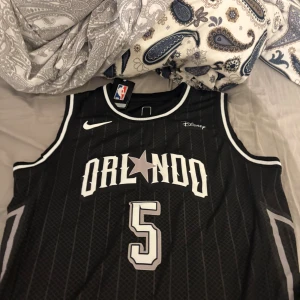Orlando Magic Banchero basketlinne - Säljer ett svart basketlinne från Nike med Orlando Magic-tryck och nummer 5, Banchero på ryggen. Linnet har vita och grå detaljer, tunna ränder och är ärmlöst. Perfekt för basket eller streetwear. Officiell NBA-logga och Disney-märke på bröstet.