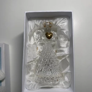 Glasängel med guldhjärta - En dekorativ glasängel med ett skimrande guldhjärta och glittriga detaljer. Ängeln har vackra vingar och en guldig gloria, perfekt som prydnad eller present. Levereras i en fin ask med satinliknande inredning.