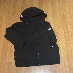 Svart Moncler vindjacka med huva - Svart vindjacka från Moncler med huva och snygga röd-vita detaljer på luvan. Jackan har dragkedja framtill, elastiska muddar och Moncler-logga på ärmen. Perfekt för blåsiga dagar och enkel att matcha med allt. Materialet är lätt och känns sportigt.