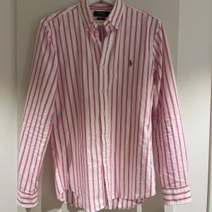 Rosa och vit randig skjorta Polo Ralph Lauren - Snygg slim fit skjorta från Polo Ralph Lauren med vertikala rosa och vita ränder. Klassisk button-down krage, långärmad och med broderad logga på bröstet. Skjortan är gjord i mjuk bomull och har knappar framtill. Perfekt för dig som gillar stilrena och fräscha plagg. Skjortan är endast prövad, iprincip helt ny!