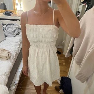 Vit kort sommarklänning med smock - Supersöt vit kort klänning med smala axelband och smockad överdel. Kjolen är lätt utställd och tyget har diskreta prickar som ger en fin struktur. Perfekt för varma dagar och enkel att styla med sneakers eller sandaler.