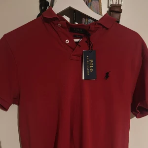 Röd pikétröja från Polo Ralph Lauren - Snygg röd pikétröja från Polo Ralph Lauren med klassisk krage och korta ärmar. Tröjan har den ikoniska broderade loggan på bröstet och är gjord i mjuk bomull. Perfekt för en stilren och avslappnad look.