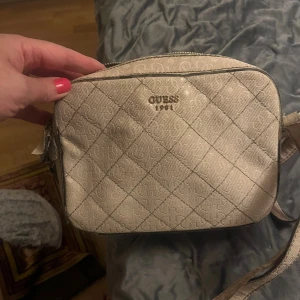 Beige axelväska från Guess - Säljer en beige axelväska från Guess med logomönster och rutigt quiltad framsida. Väskan har justerbar axelrem och dragkedja upptill. Perfekt storlek för det viktigaste och snygg detalj till din outfit.