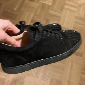 Svarta sneakers med nitar och röd sula - Säljer ett par svarta sneakers i mocka med coola svarta nitar över tån och klassisk röd sula undertill. Skorna har snörning och en platt sula, vilket ger en edgy vibe. Perfekta för dig som vill sticka ut med din stil.